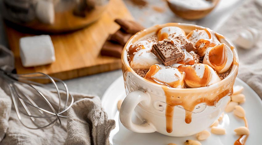 Biela šálka plná horúceho čokoládového nápoja s karamelom a marshmallows