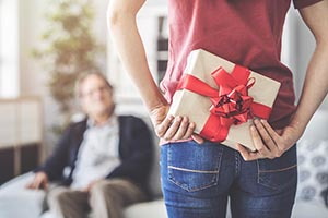 Hľadáte darček pre frfloša? Vtipné a netradičné vianočné darčeky podľa povahy človeka Žena, stojaca pred mužom sediacim na pohovke, pre neho za chrbtom skrýva zabalený darček s červenou mašľou